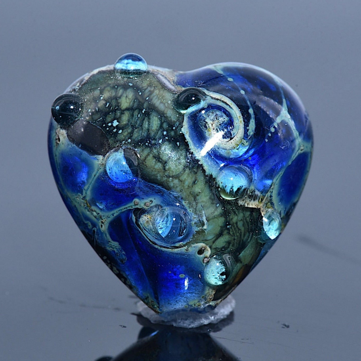 Heart-shaped blue glass bead on a reflective surface with a gray background