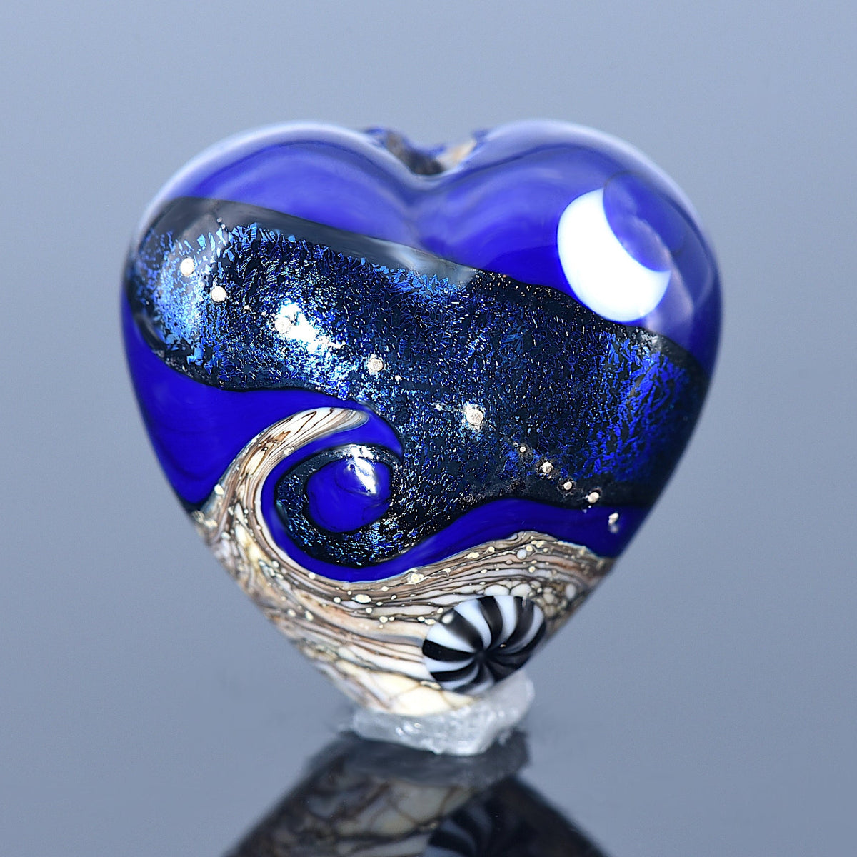 Heart-shaped glass sculpture with blue and silver design on a reflective surface