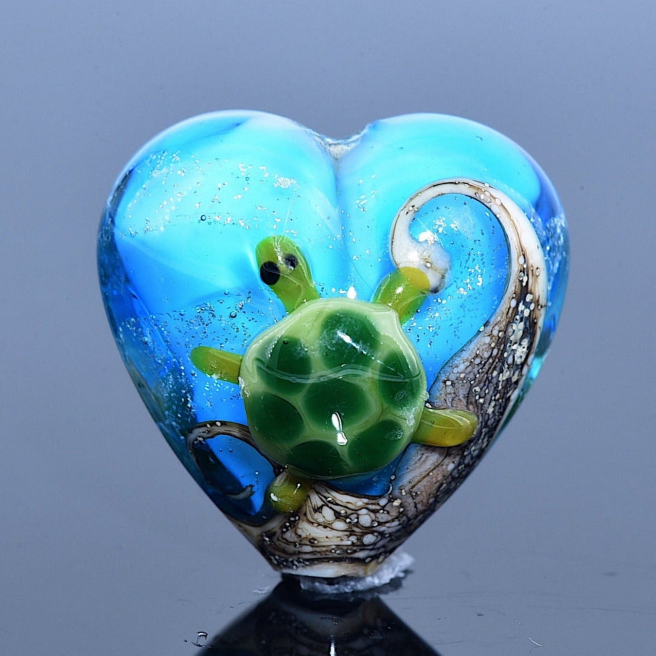 Heart-shaped blue glass ornament with a green turtle and brown seaweed on a reflective surface.