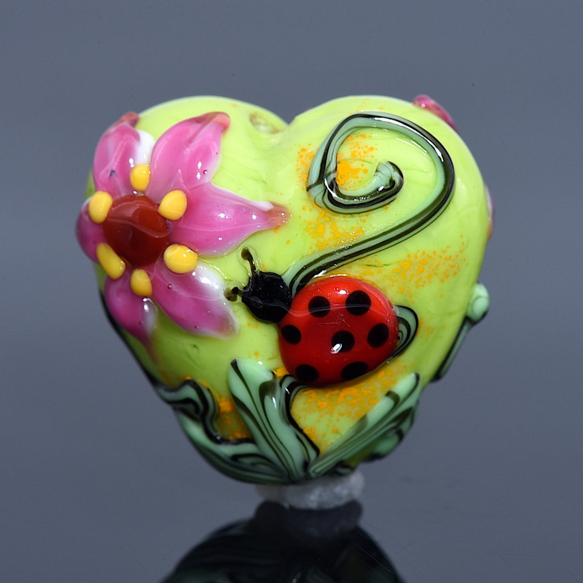 Colorful heart-shaped object with floral design and ladybug on a reflective surface