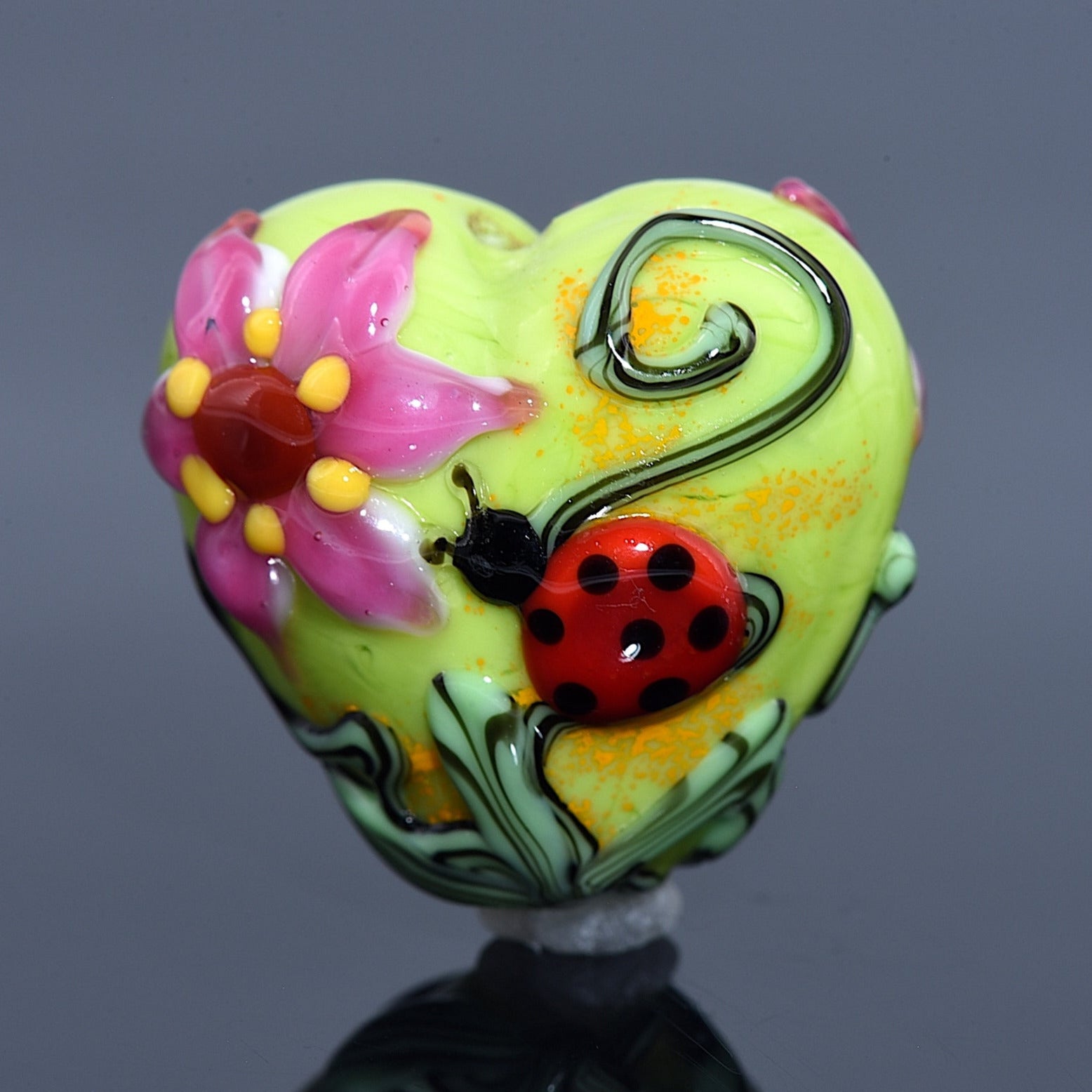 Colorful heart-shaped object with floral design and ladybug on a reflective surface